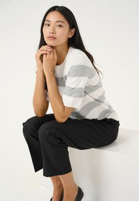 Gestreifter Pullover in Weiß und Grau, Baumwollmischung, kombiniert mit schwarzen, verkürzten Hosen. Modell sitzt, Hände verschränkt, mit langen dunklen Haaren.