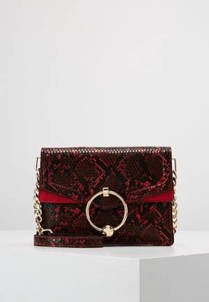 Petit sac rectangulaire rouge et noir à motif peau de serpent, avec anneau doré et bandoulière chaîne, posé sur une surface blanche.