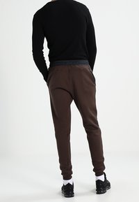 Pantalones de chándal marrones con un diseño ajustado, cintura elástica y tejido texturizado. Llevados con zapatillas negras y calcetines blancos.