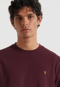T-shirt in cotone bordeaux con scollatura a girocollo e maniche corte. Presenta un piccolo logo giallo ricamato sul lato sinistro del petto.