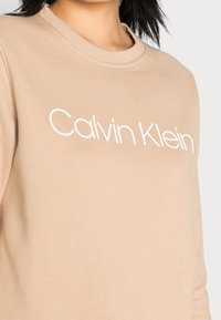 Sudadera beige hecha de tejido suave, con un cuello redondo y el nombre de la marca "Calvin Klein" impreso en blanco en la parte frontal.