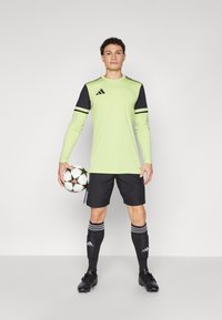 Groen sportshirt met lange mouwen en zwarte accenten, zwarte shorts, zwarte sokken met witte strepen en zwarte voetbalschoenen, terwijl hij een voetbal vasthoudt.