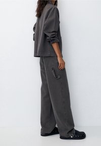 Giacca di denim grigia con spalle basse, abbinata a pantaloni grigi a gamba larga con tasca laterale e risvolti. Calzature nere con accenti argentati.