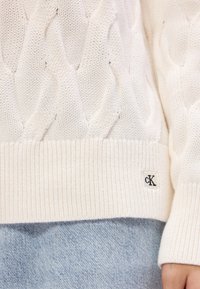 Biely pletený sveter s káblovým vzorom a rebrovaným lemom, na ktorom je malý čierny štítok s logom "cK". Nosený cez svetlo modré denimové nohavice.