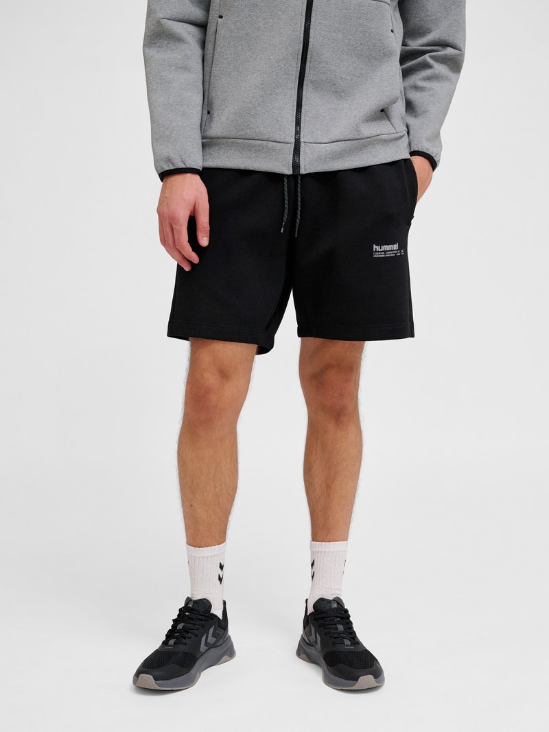 Homme portant une veste grise zippée, un short noir avec logo, des chaussettes blanches et des baskets noires, debout devant un fond uni.