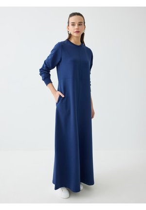 Frau mit dunklen Haaren, die ein langes dunkelblaues Kleid und weiße Sneaker trägt, steht mit einer Hand in der Tasche vor einem schlichten Hintergrund.
