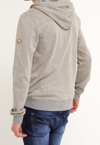 Felpa grigia con cappuccio in tessuto testurizzato, zip frontali e polsini a costine. Presenta una toppa con logo sulla manica sinistra e una vestibilità comoda.