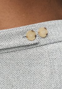 Υφασμα σε ανοιχτό γκρι με σχέδιο ψαροκόκκαλο και δύο στρογγυλά μπεζ κουμπιά ραμμένα κοντά σε διπλωμένη άκρη.