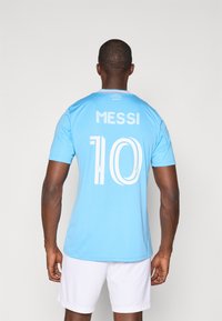 Camisola desportiva azul clara com o nome "MESSI" e o número "10" em branco nas costas. Mangas curtas, textura lisa e gola redonda.