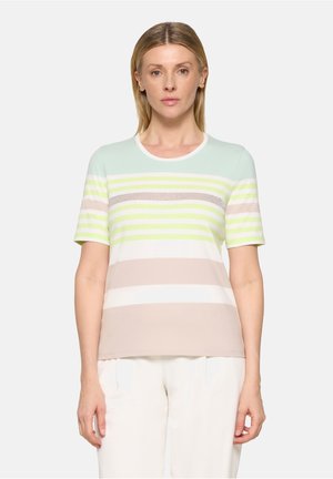 Femme aux cheveux blonds raides portant une chemise à manches courtes à rayures pastel et un pantalon blanc, face à l'avant sur un fond uni.