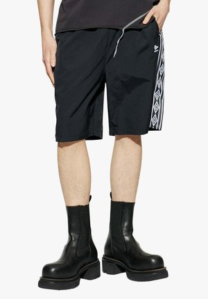 Persona che indossa pantaloncini neri con motivo a coulisse e stivali alla caviglia in pelle nera, in piedi davanti a uno sfondo bianco semplice.