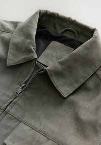Chaqueta de ante en verde oliva con cuello plano, cierre de cremallera y dos bolsillos frontales. El interior cuenta con un forro negro suave.