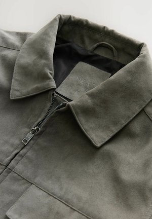 Olivengrüne Wildlederjacke mit flachem Kragen, Reißverschluss und zwei Vordertaschen. Das Innere hat ein glattes schwarzes Futtermaterial.