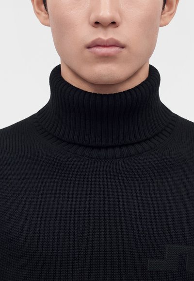 Gros plan du bas du visage et du cou d'une personne portant un pull à col roulé côtelé noir avec un logo discret sur la poitrine.