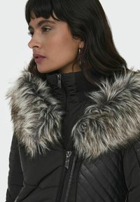 Sort pufferjakke med en aftagelig grå faux fur-krave, der har en frontlynlås og quiltet tekstur på kroppen.