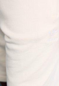 Kreminės spalvos Adidas sportinės kelnės su raukšlėta tekstūra, ant kišenės yra įspausta logo. Prigludusi dizaino laikysena, su lygiu paviršiumi.