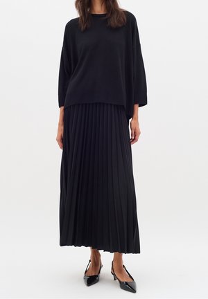 Kobieta ubrana w luźną czarną bluzkę z długim rękawem, czarną plisowaną spódnicę midi oraz czarne czółenka typu slingback z ostrym noskiem, stojąca na białym tle.