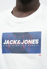 Λευκό βαμβακερό t-shirt με ορθογώνιο σχέδιο σε μπλε χρώμα με κάθετες ρίγες και έντονο λευκό κείμενο «JACK & JONES», με κόκκινες λεπτομέρειες.