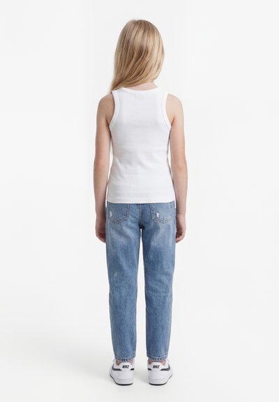 Bērns ar gariem blondiem matiem, tērpts baltā bez piedurknēm kreklā, zilās džinsu biksēs un baltās Nike sporta kurpēs, stāvot ar muguru pret baltu fonu.