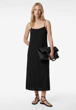 Femme en robe midi noire à bretelles spaghetti tenant une grande pochette noire tissée, portant des sandales plates noires cloutées, debout sur fond blanc.