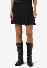 Črna plisejska mini krilo z gladko teksturo tkanine, v kombinaciji z visokimi črnimi usnjenimi škornji z zaponkami.