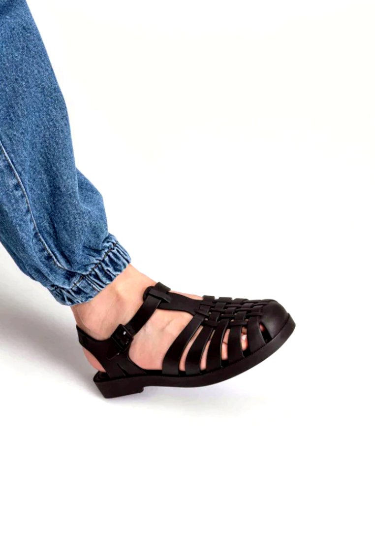 Sandali neri in gomma con design intrecciato, open-toe e cinturino alla caviglia regolabile. Tacco arrotondato e suola testurizzata. Indossati con joggers in denim blu.