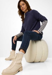 Kvinde sidder på en rund, tekstureret skammel iført en mørkeblå sweater over stribede lange ærmer, mørke jeans og beige ankelstøvler med kraftige såler.