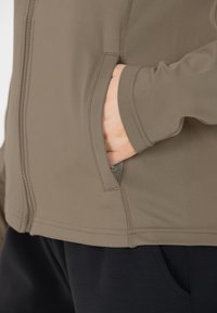 Leichte, taupefarbene Zip-Jacke mit glatter Textur. Verfügt über Seitentaschen und kontrastierende Nähte, kombiniert mit schwarzen Hosen.