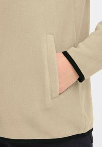 Beige fleecegenser med sidelomme. Har svart kant rundt ermene og nederkanten. Myk tekstur og avslappet passform.