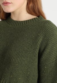Sweter robiony na drutach w ciemnozielonym kolorze z żebrowanym dekoltem. Posiada teksturowane body z drobnymi wtrąceniami jaśniejszych kolorów.