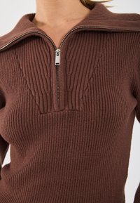 Pull marron côtelé avec un col montant et une demi-fermeture éclair. Présente des détails texturés le long du col et du panneau avant.