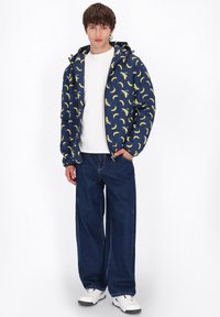 Giacca blu navy con stampa di banane gialle, chiusura con zip e cappuccio, abbinata a jeans scuri a vestibilità ampia e sneakers bianche.