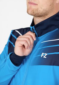 Blå sportjacka med mörkblå accenter, dragkedja i kragen och vita ränder. Materialet verkar vara slätt; har en logotyp på bröstet.