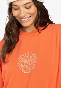 Oranje sweatshirt van zacht katoen, met een ronde afbeelding van "Sinds 1973" en een palmboomontwerp in lichtgrijs op de borst.