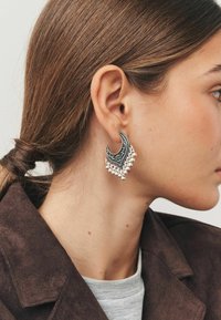 Sølvsmykke formet som en halvmåne med detaljeret udsmykning og hængende perleakcenter. Designet med en reflekterende overflade.