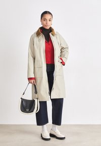 Beige raincoat with a brown collar, red button-up sweater underneath, dark jeans, white socks, and black handbag with faux fur accents.
