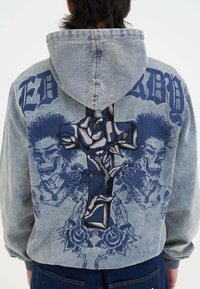 Sudadera de mezclilla con un tono azul grisáceo. Presenta una gran cruz bordada con una rosa, gráficos de calaveras y texto en negrita en la parte posterior.