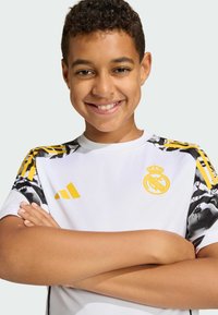 Hvid sportstrøje med gule detaljer, sorte og hvide mønstrede skuldre og et rundt emblem på brystet. Designet til børn.