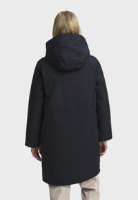 Personne aux cheveux blonds portant un long manteau d'hiver noir à capuche et un pantalon beige, se tenant de dos contre un fond uni.