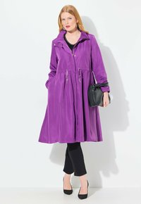Manteau léger violet avec taille à cordon, manches longues et devant boutonné. Associé à un pantalon noir et des talons, avec un petit sac noir.