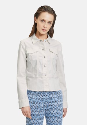 Giacca di jeans bianca con chiusura a bottoni, due tasche sul petto e un design leggermente aderente, abbinata a pantaloni blu a fantasia.