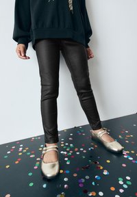 Pantalones ajustados de cuero sintético negro combinados con zapatos Mary Jane metálicos dorados que cuentan con dos correas, sobre un fondo de colorido confeti.
