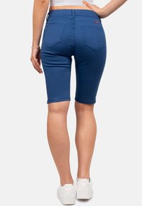 Bermuda shorts in denim blu con un design aderente, tasche posteriori e orli risvoltati. Abbinati a sneaker bianche.
