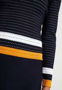Dettaglio ravvicinato di un maglione nero a coste con motivo a pois bianchi, caratterizzato da strisce orizzontali di colore bianco e arancione in contrasto sul punto vita e sui polsini delle maniche.