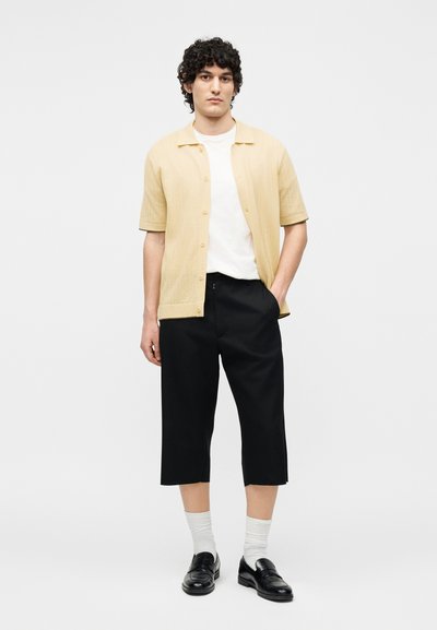 Νέος άνδρας με σγουρά μαλλιά, φορώντας μπεζ κοντομάνικο πουκάμισο με κουμπιά, λευκό μπλουζάκι, μαύρο φαρδύ κομμένο παντελόνι, λευκές κάλτσες και μαύρα μοκασίνια.