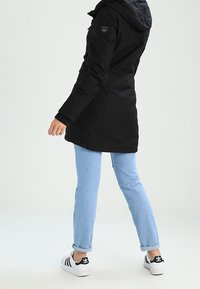 Svart huva parkas med en slät textur, sidfickor och en logotyp på ärmen, i kombination med ljusblå jeans och svarta sneakers.