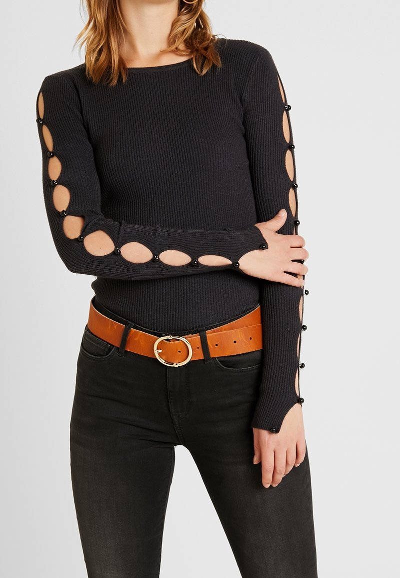 Femme portant un pull noir à côtes avec des découpes ovales et des perles noires le long des manches, associé à un jean noir et une ceinture en cuir marron.