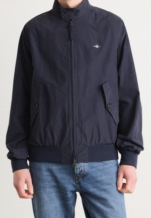 Homme portant une veste bleu marine zippée avec col boutonné et poches, associée à un jean bleu, visible du cou aux hanches.