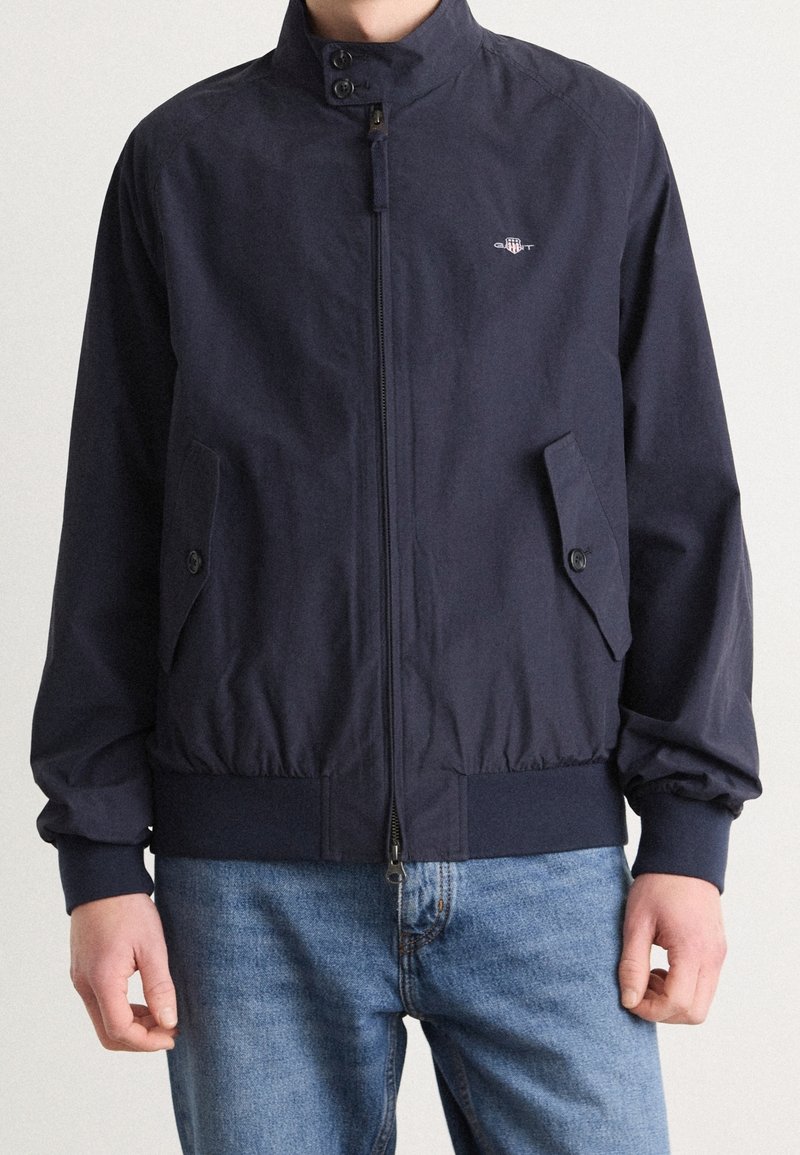 Homme portant une veste bleu marine zippée avec col boutonné et poches, associée à un jean bleu, visible du cou aux hanches.