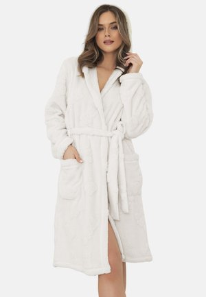 Kobieta ubrana w puszysty, biały szlafrok z kieszeniami i paskiem, stojąca z ręką przy twarzy i zrelaksowanym wyrazem twarzy.